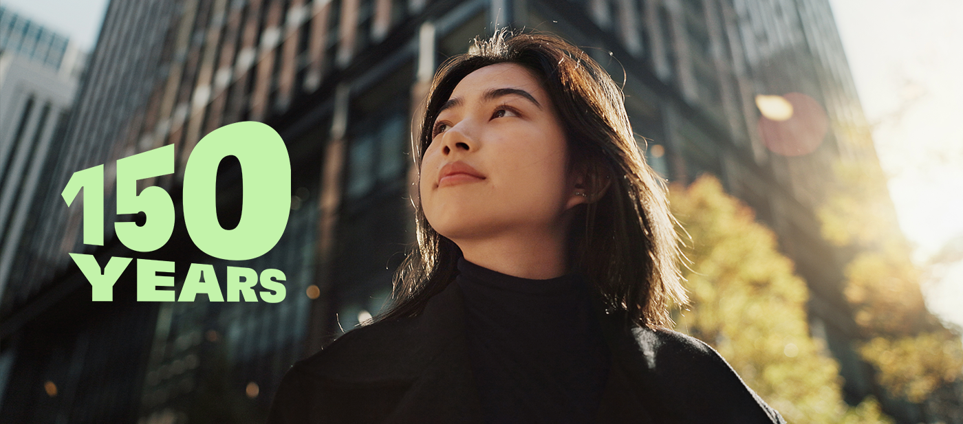 ガラス張りの高層ビルと秋らしい樹木が並ぶ現代的な都市の景観に「150 YEARS」のグリーンの文字を配した、創業150周年の祝賀を象徴する画像。