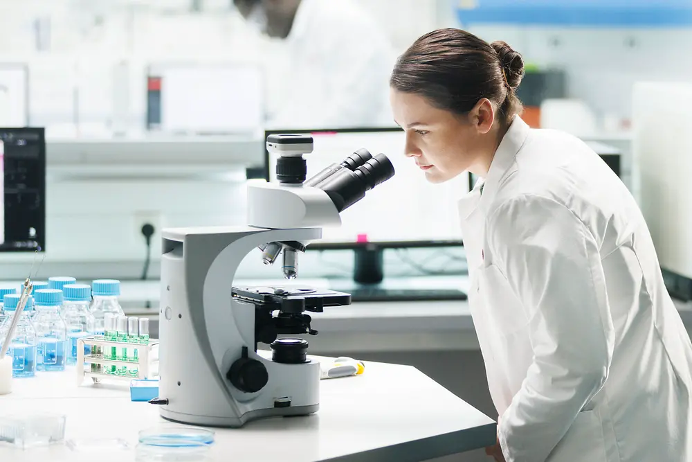A woman in a white lab coat is leaning over a microscope in a laboratory. On the table in front of her, there are several vials and laboratory utensils. In the background, another person in a white lab coat is working on a computer.