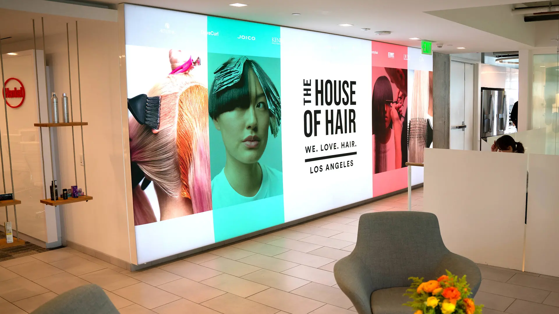 近代的なロビーの明るい壁に、カラフルな髪をテーマにしたイラストと「THE HOUSE OF HAIR – WE.LOVE.HAIR. – LOS ANGELES」の文字が書かれた複数の大きなパネルが掛かっている様子。タイル張りの床と白いパーテーションによるミニマルなデザインが採用された空間に、ライトグレーのラウンジチェアーと鮮やかなフラワーアレンジメントを乗せた小さなテーブルが置かれている。