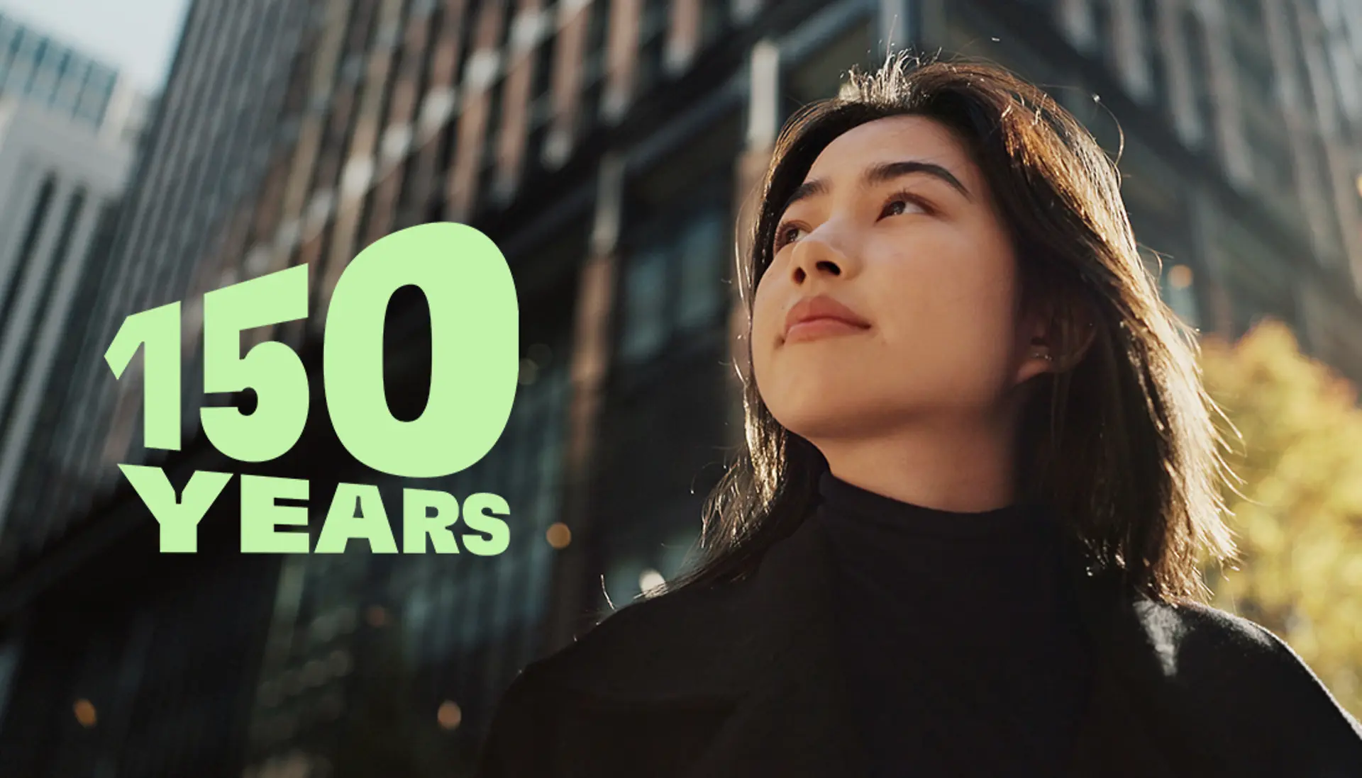 ガラス張りの高層ビルと秋らしい樹木が並ぶ現代的な都市の景観に「150 YEARS」のグリーンの文字を配した、創業150周年の祝賀を象徴する画像。