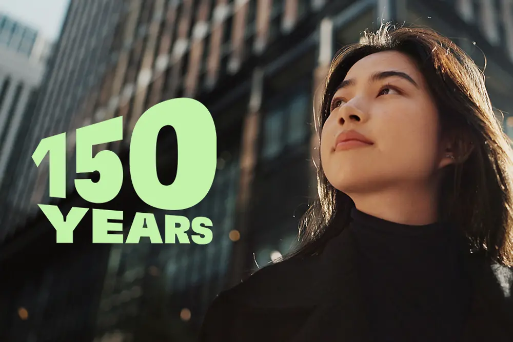 ガラス張りの高層ビルと秋らしい樹木が並ぶ現代的な都市の景観に「150 YEARS」のグリーンの文字を配した、創業150周年の祝賀を象徴する画像。