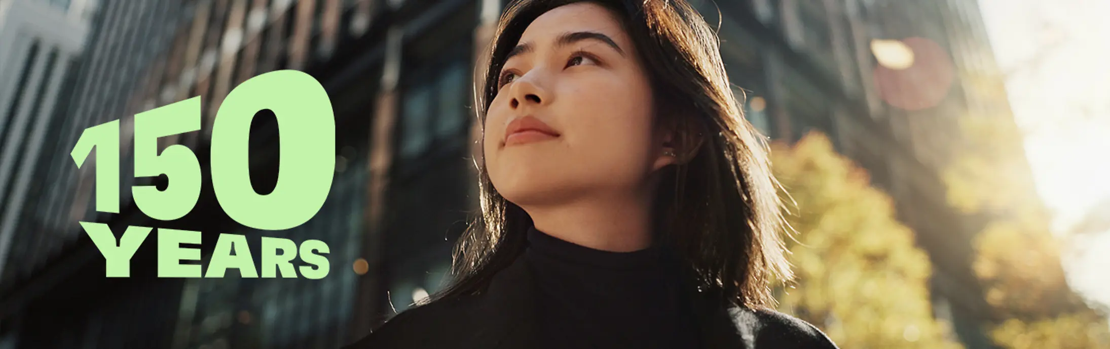 ガラス張りの高層ビルと秋らしい樹木が並ぶ現代的な都市の景観に「150 YEARS」のグリーンの文字を配した、創業150周年の祝賀を象徴する画像。